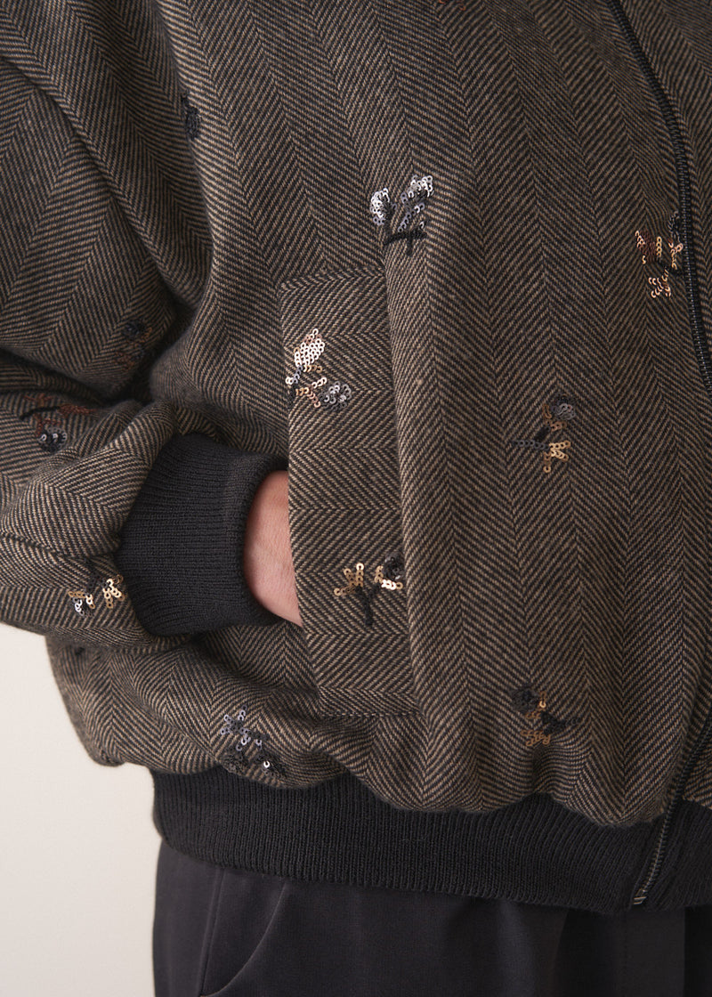 Close up of pocket detail on person wearing a brown textured bomber jacket with sequin detail and black trousers on a plain background