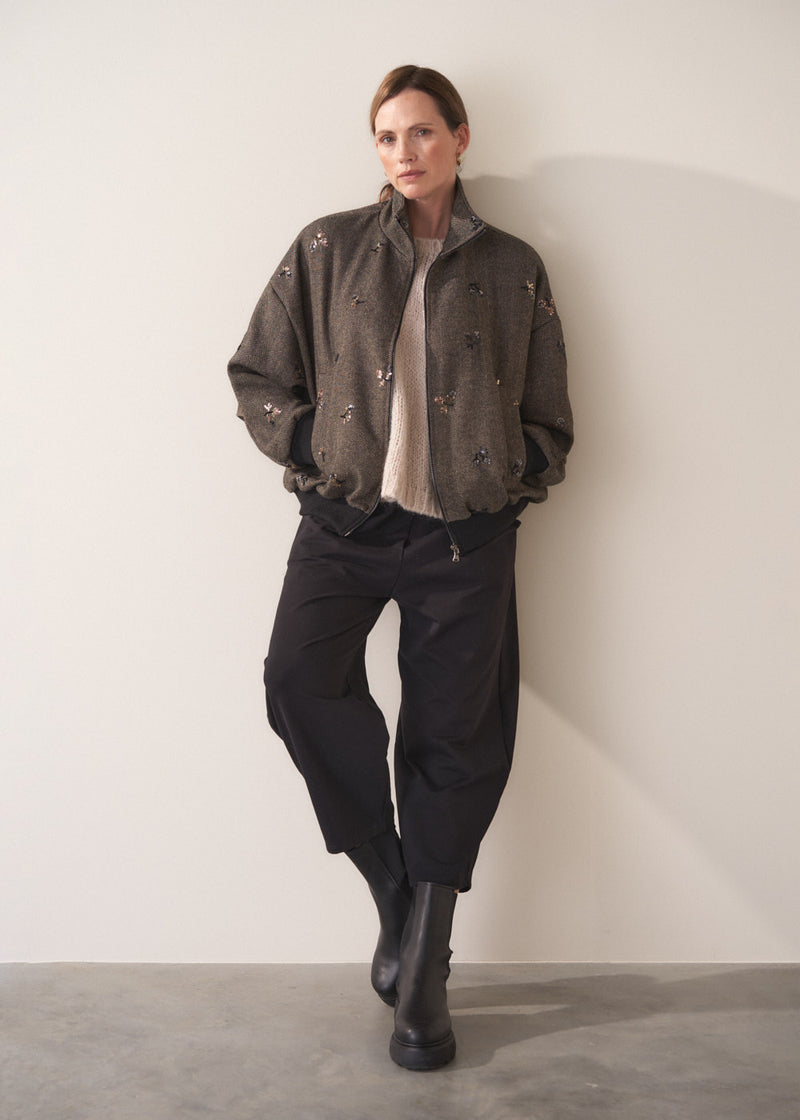 Person leaning against a beige background wearing a brown textured bomber jacket with sequin detail and black trousers