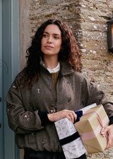 A woman in dark jeans and a brown, textured bomber jacket, featuring sequin detailing, holding wrapped presents