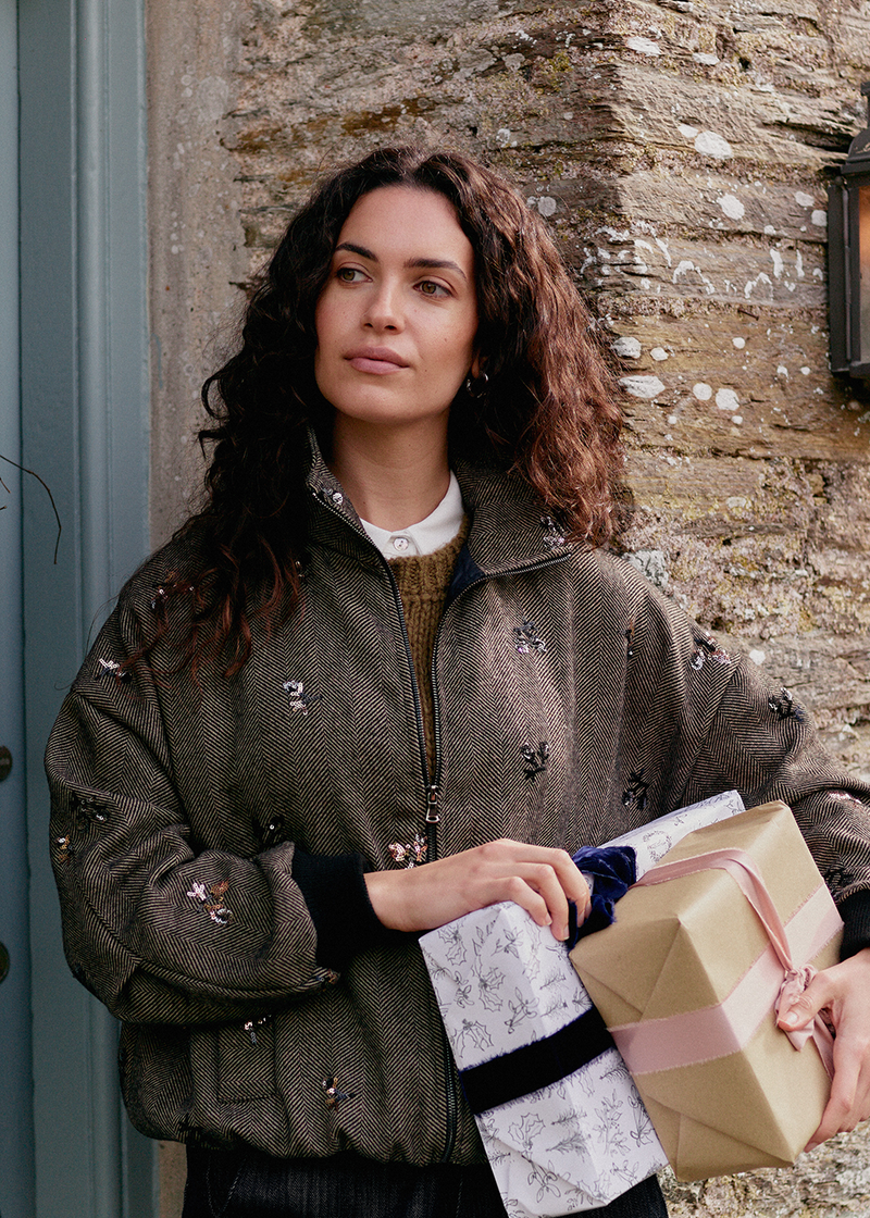 A woman in dark jeans and a brown, textured bomber jacket, featuring sequin detailing, holding wrapped presents