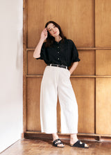 A woman model wearing white cropped barrel-leg jeans, a black top, and sandals, against a wooden background.