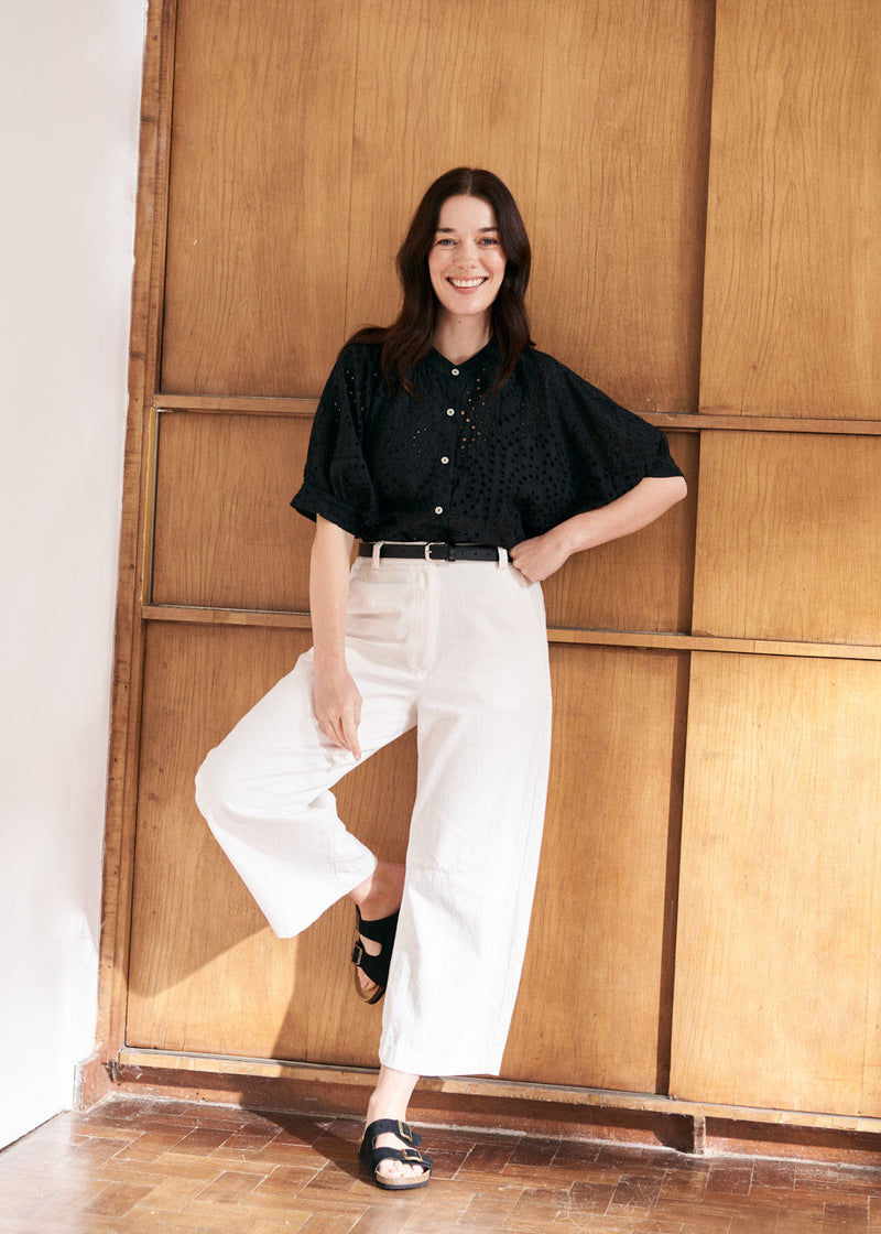 Model wearing a black broderie anglaise top with belted white wide leg trousers and black sandals, against a natural wood background