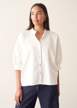 A woman wearing a white-button down cotton shirt with three-quarter puff sleeves and a relaxed fit, with blue denim jeans.