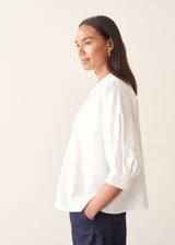 A woman wearing a white-button down cotton shirt with three-quarter puff sleeves and a relaxed fit, with blue denim jeans.