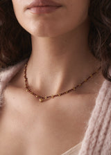 Close up of woman wearing a necklace with plum crystal beads, gold discs, and a hammered heart pendant, with pink chunky knit cardigan