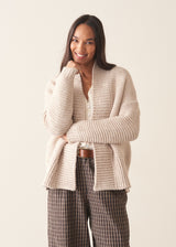 A woman wearing a chunky knit open front cardigan in oatmeal beige with checked barrel leg trousers