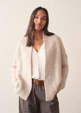 A woman wearing a chunky knit open front cardigan in oatmeal beige with checked barrel leg trousers