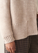 A woman wearing a chunky knit open front cardigan in oatmeal beige with checked barrel leg trousers