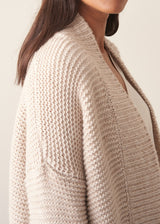 A woman wearing a chunky knit open front cardigan in oatmeal beige with checked barrel leg trousers