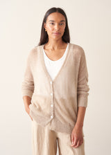 A woman wearing a soft knit cardigan in a light beige colour, worn over a white vest top and paired with matching beige trousers.