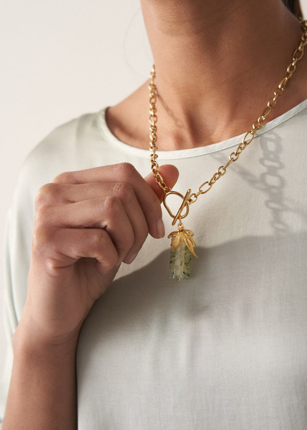 Woman in a light blue top wearing and holding a chunky gold chain necklace with a sage crystal pendant