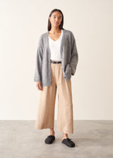 A woman wearing a grey knit cardigan over a white top and beige trousers, photographed against a plain studio background.