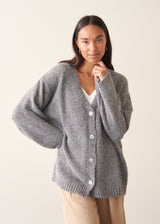 A woman wearing a grey knit cardigan over a white top and beige trousers, photographed against a plain studio background.