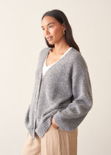 A woman wearing a grey knit cardigan over a white top and beige trousers, photographed against a plain studio background.