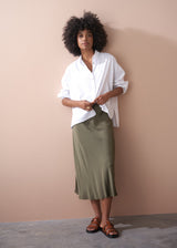 Woman leans against a wall wearing a white oversized shirt with green satin skirt and sandals