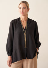 A woman modeling a black long-sleeve blouse with a split neckline and a decorative beaded tassel necklace, paired with beige pants, against a plain beige background.