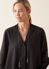 A woman wearing a black long-sleeve blouse and layered silver necklaces, photographed against a plain light beige studio background.