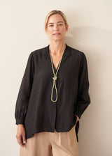 A woman wearing a black long-sleeve button-up blouse with a split neckline and a long knotted beaded necklace, paired with beige trousers, standing against a plain light beige wall.