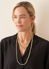 A woman with blonde hair tied back, wearing a black textured blouse and a long, double-strand light green beaded necklace, photographed against a plain light beige background.