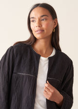A woman wearing a black embroidered textured cropped jacket over a white top and dark blue jeans.
