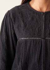 A woman wearing a black embroidered textured cropped jacket over a white top and dark blue jeans.