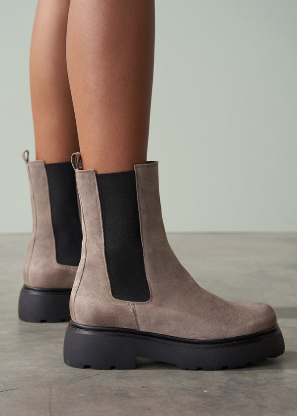 Brown suede boots with black soles worn by a person on a neutral background