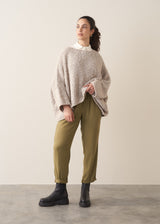 Woman wearing a creamy white blouse, boucle jumper and green tapered trousers against a plain background