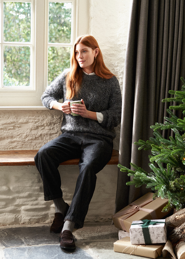 Model relaxing in a window seat wearing a grey sweater, paired with blue black cotton trousers and brown suede loafers