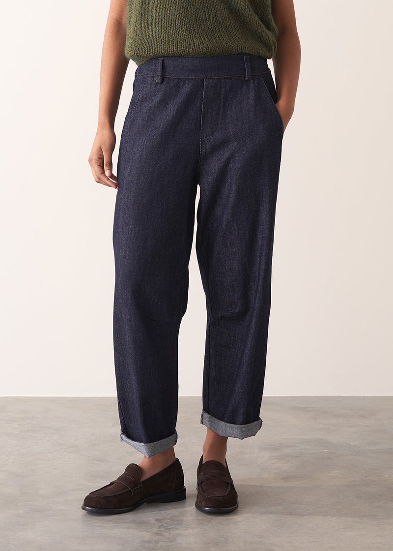 Woman wearing a green sleeveless sweater and dark blue jeans against a plain background