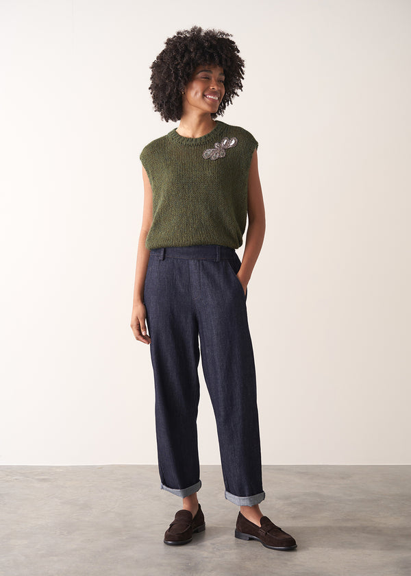 Woman wearing a green sleeveless sweater and dark blue jeans against a plain background