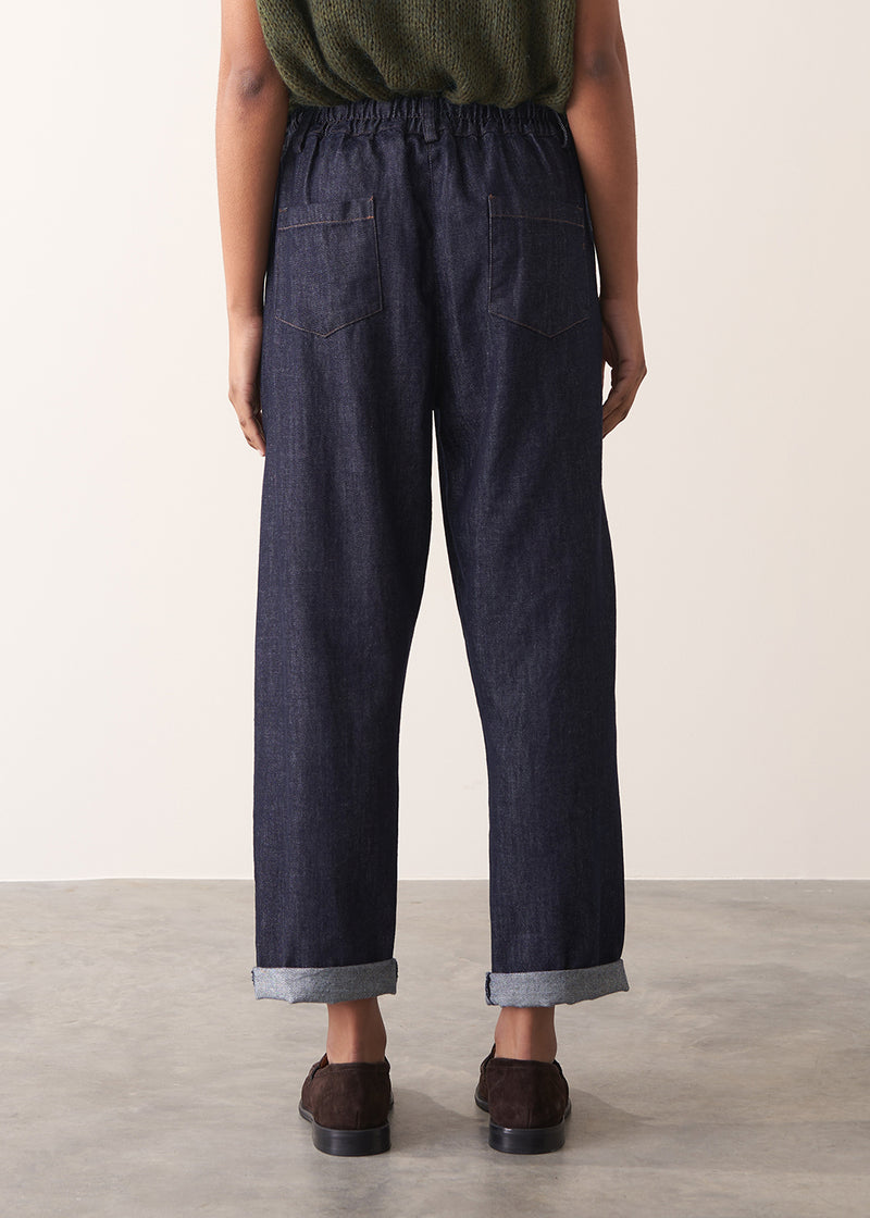 Woman wearing a green sleeveless sweater and dark blue jeans against a plain background