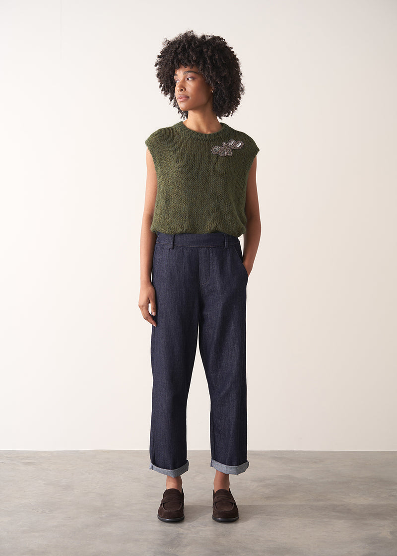 Woman wearing a green sleeveless sweater and dark blue jeans against a plain background