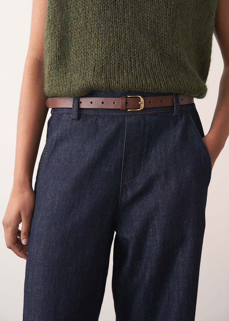 Woman wearing a green sleeveless sweater and dark blue jeans against a plain background