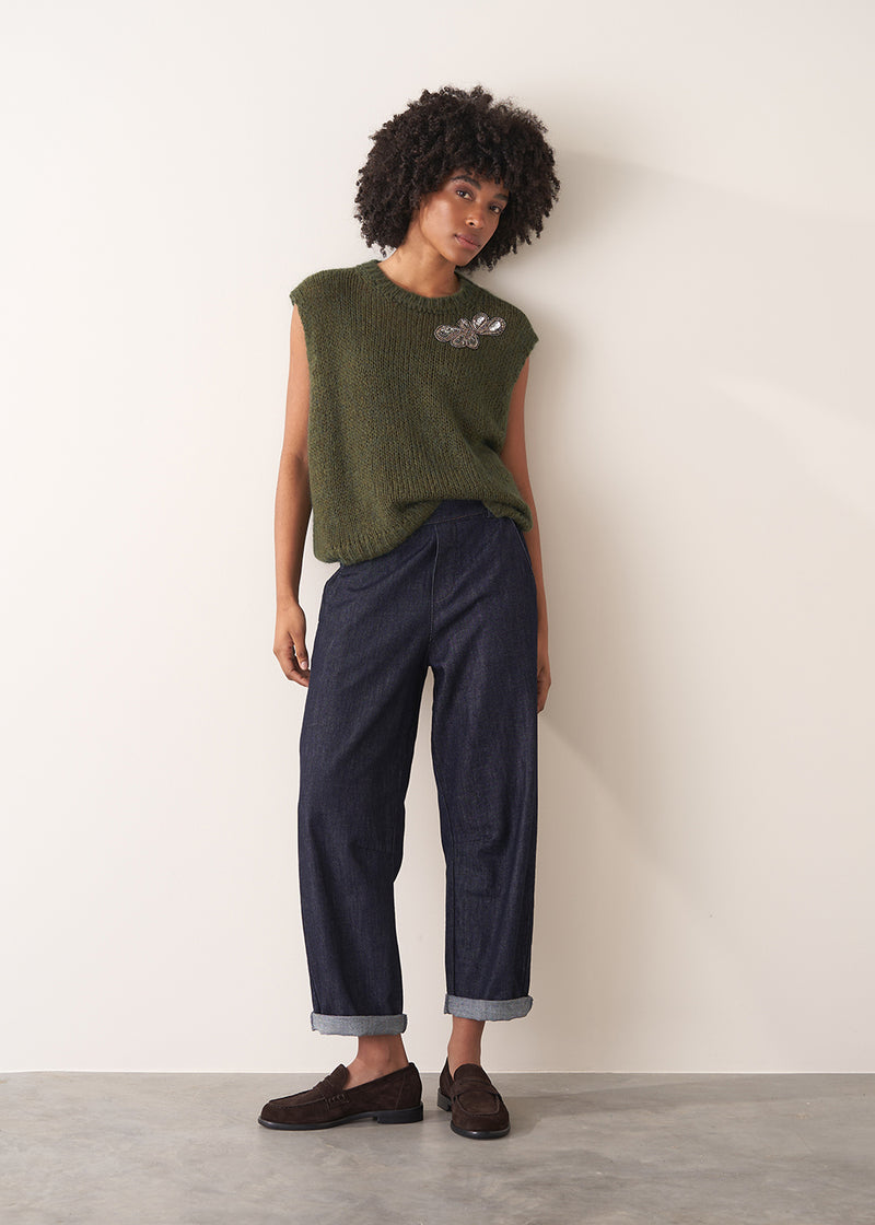 Woman wearing a green sleeveless sweater and dark blue jeans against a plain background