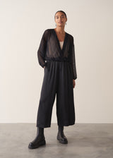 Women wearing a long sleeve sheer sparkle draped top with cream satin camisole and black satin trousers, against a beige background