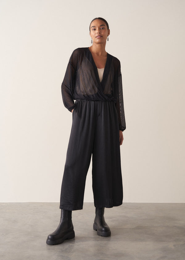 Women wearing a long sleeve sheer sparkle draped top with cream satin camisole and black satin trousers, against a beige background