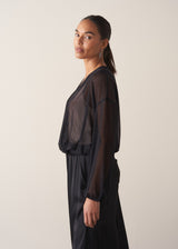 Women wearing a long sleeve sheer sparkle draped top with cream satin camisole and black satin trousers, with a beige background 