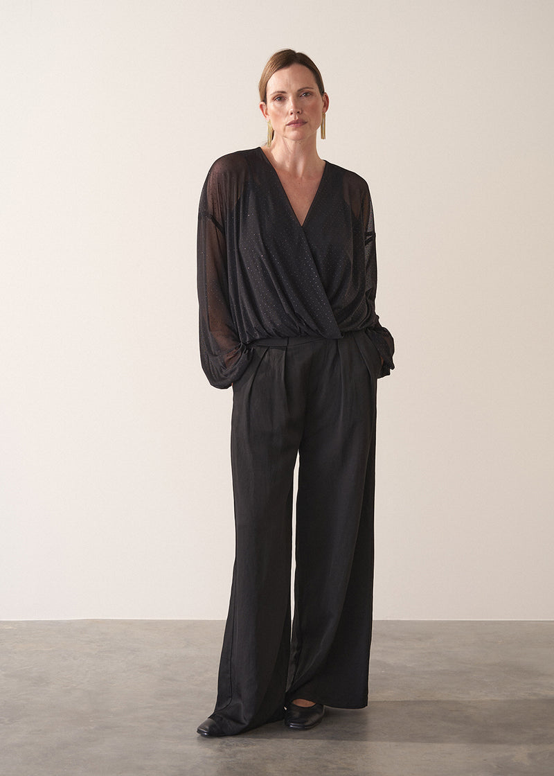 Woman wearing a black sheer top and black wide leg trousers against a plain background