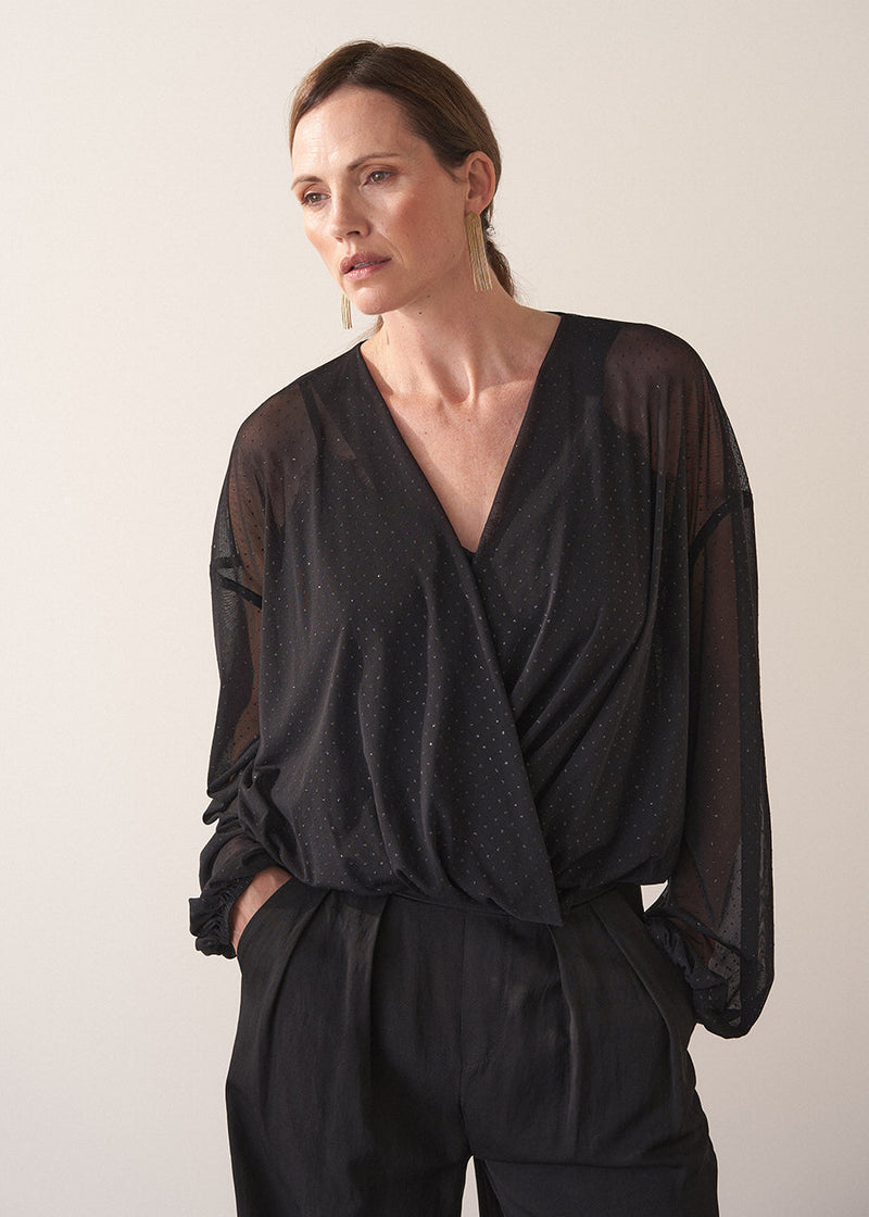 Woman wearing a black sheer top and black wide leg trousers against a plain background