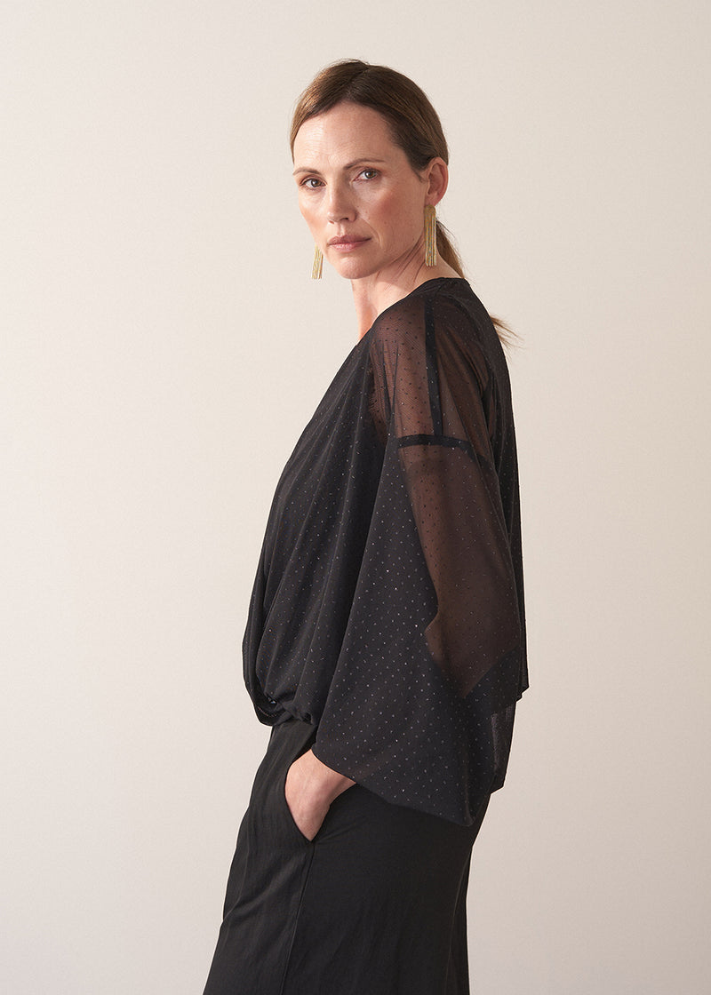 Woman wearing a black sheer top and black wide leg trousers against a plain background