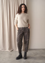 Woman stood against a fabric backdrop wearing taupe leopard print barrel leg trousers with an oatmeal sweater and black chunky leather boots