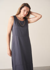 A woman wearing a sleeveless, maxi-length dark blue grey dress with a cocoon skirt