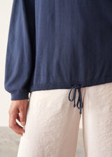 A woman wearing a navy blue long-sleeve top with a wide boat neckline and front drawstring hem, paired with light beige trousers.