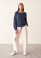 A woman wearing a navy blue long-sleeve top with a wide boat neckline and front drawstring hem, paired with light beige trousers.