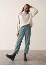 Woman wearing a cream rollneck sweater and aqua cord trousers against a plain background