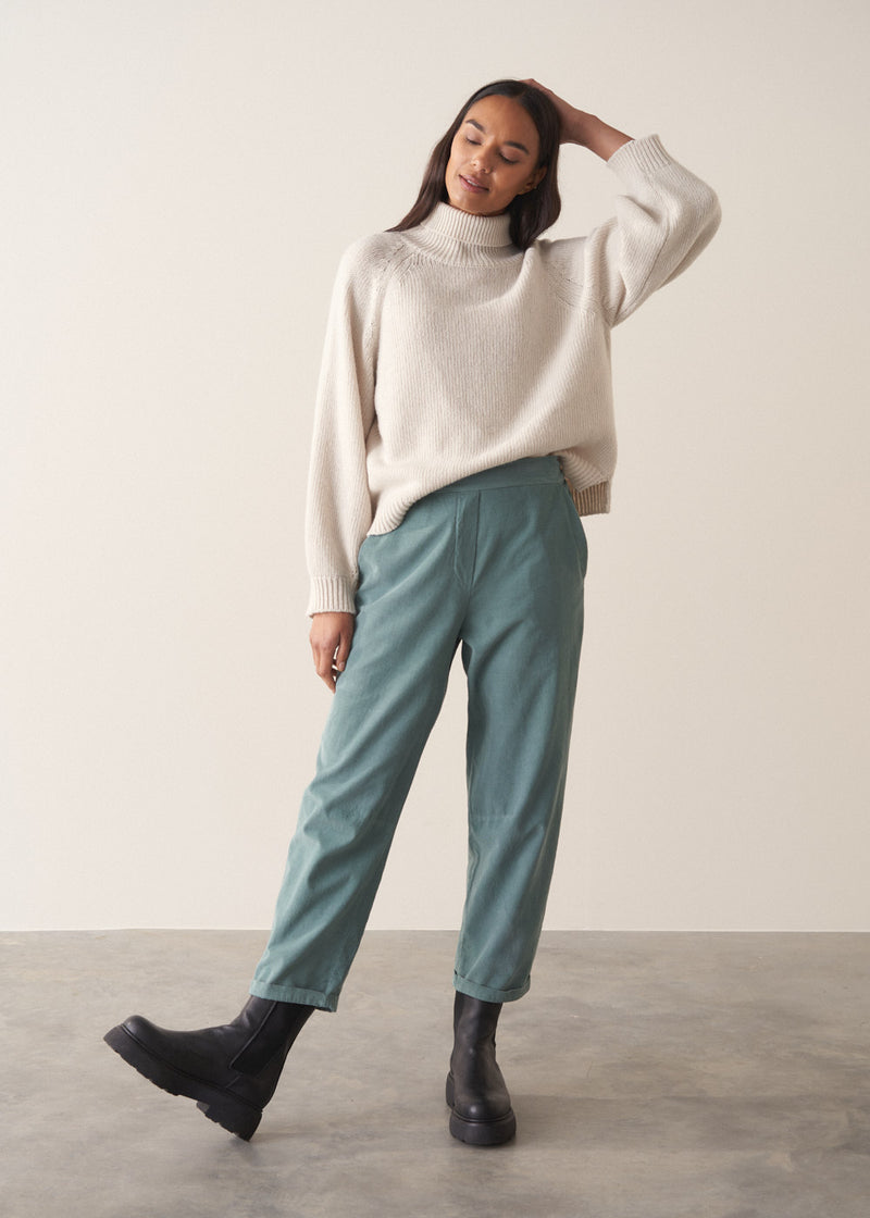 Woman wearing a cream rollneck sweater and aqua cord trousers against a plain background