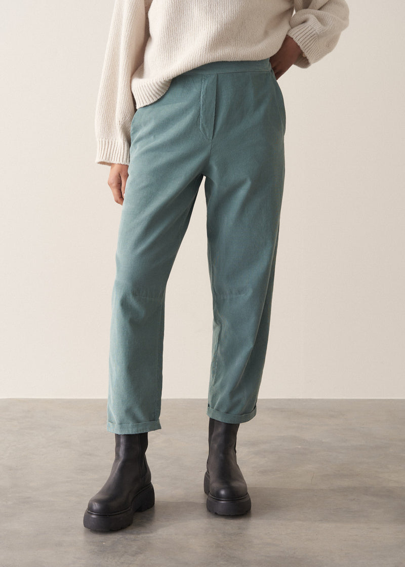 Woman wearing a cream rollneck sweater and aqua cord trousers against a plain background