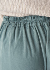 Woman wearing a cream roll neck sweater and aqua cord trousers against a plain background