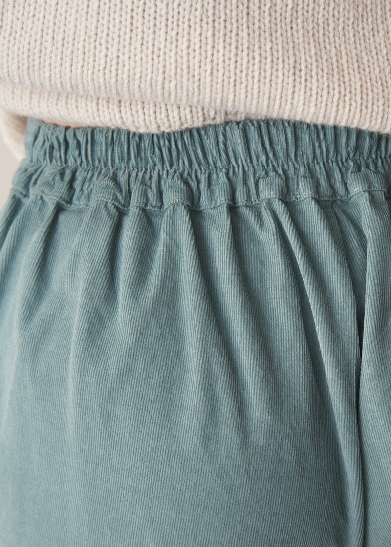 Woman wearing a cream roll neck sweater and aqua cord trousers against a plain background