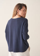 A woman stands against a plain off-white wall wearing a navy blue knit top with a brown tie detail, white wide-leg trousers, and beige slip-on shoes.